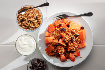 A breakfast with papaya granola and blackberries on a white plate and on a white table with a small...