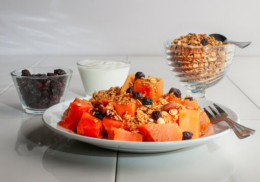 A Breakfast With Papaya Granola And Blackberries On A White Plate And On A White Table With A Small Cup Of Yogurt, Granola And Blackberries On The Side, Side View Shot.