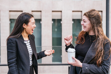 Asian woman in formal suit joyfully laughing while talking to colleague standing with reusable thermo tumbler during break in work