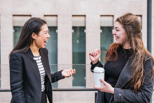 Asian woman in formal suit joyfully laughing while talking to colleague standing with reusable thermo tumbler during break in work