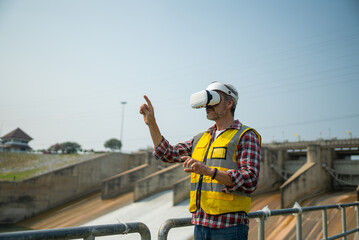 Portrait of engineer wearing yellow vest and white helmet using augmented reality application in virtual reality glasses. Working on a dam with a hydroelectric power plant. Renewable energy systems