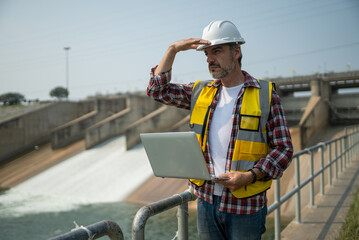 Naklejka na ściany i meble Portrait of engineer wearing yellow vest and white helmet with laptop computer Working day on a water dam with a hydroelectric power plant. Renewable energy systems, Sustainable energy concept