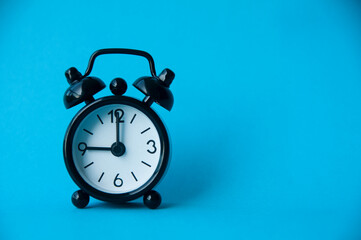 Black alarm clock isolated on light blue background. The clock set at 9 o'clock.