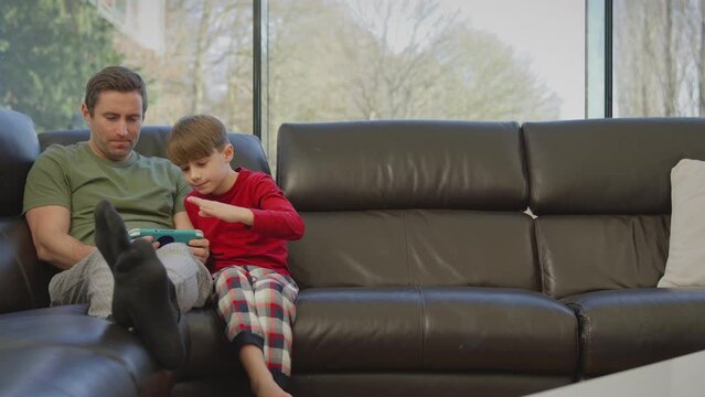 Father with son wearing pyjamas sitting on sofa at home playing computer game on handheld device - shot in slow motion