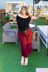 A fashionable girl with curvy curves in red bell-bottomed trousers and a black T-shirt dances happily laughing and holding two glasses with cocktails in her hands on the roof of the restaurant