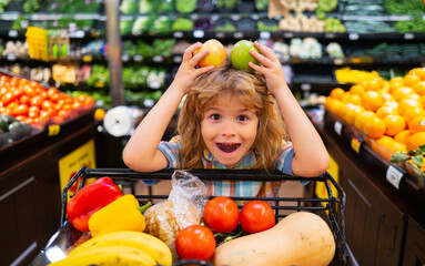 Child choosing a apple in a store. Funny little child smile and holding apple. Kid shopping in supermarket. Little boy with cart choosing fresh vegetables in local store.