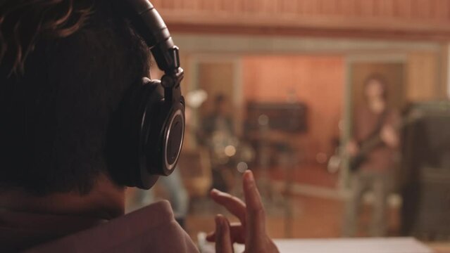 Rear view slowmo of music producer in headphones standing by mixing board at music record studio listening to rock band performing new track