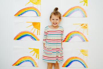 Cheerful little girl in a dress shirt next to six paintings of sun and rainbow