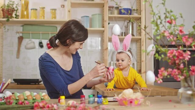 Mother with baby decorating easter eggs. Baby with bunny ears at the kitchen desk.