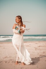 Fototapeta na wymiar Model in boho style in a white long dress and silver jewelry on the beach. Her hair is braided, and there are many bracelets on her arms.