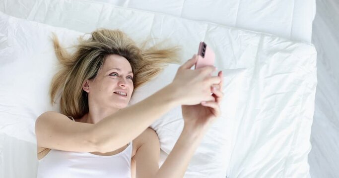 Smiling beautiful woman looks at smartphone screen and lies on bed