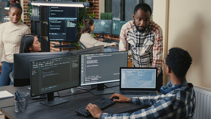 Programer sitting at desk with multiple screens running code talking with colleague about...