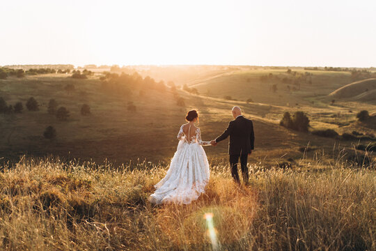 Wedding. Nature. The groom in a suit and the bride in a white dress are standing in an autumn field against the backdrop of the sunset sky and the sun's rays