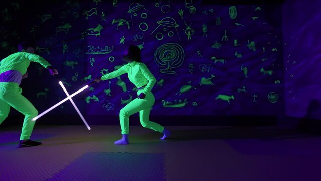 Couple of young people are having fun in a neon amusement park, guy and a female are fighting with each other using lightsabers, 4k slow motion.