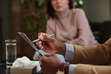 Close-up of black psychologist making notes about patient in clipboard while listening to her during therapy