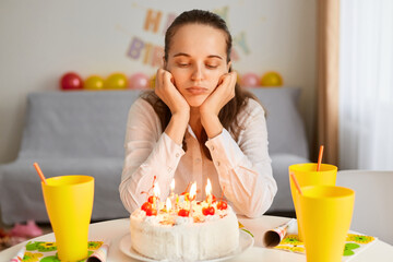 Indoor shot of bored sad Caucasian young adult woman sitting at table and looking at birthday cake, being upset to celebrate alone, waiting her guests.