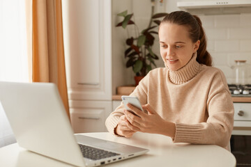 Image of delighted smiling adorable woman wearing beige jumper posing in kitchen and using cell phone, holding smart phone in hands and typing, answering messages.