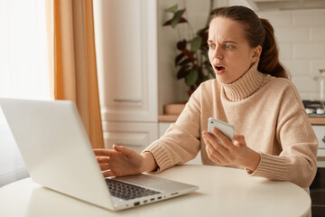 Portrait of shocked astonished woman wearing beige jumper posing in kitchen and working on laptop, holding cell phone and looking at notebook with open mouth and surprise.