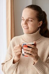 Portrait of young adult attractive woman wearing beige sweater holding cup of coffee or tea, looking away at window, dreaming about something pleasant and smiling.
