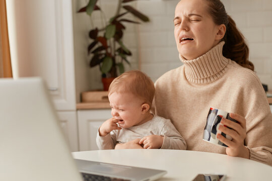 Image of crying woman wearing beige jumper posing in kitchen and working on laptop and holding baby, can't calm down child, sitting at table in front of notebook.