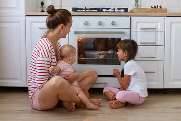 Indoor shot of happy woman with bun hairstyle wearing striped casual style shirt sitting on floor in kitchen with her children, mother and her kids waiting for tasty dessert, baking in oven.