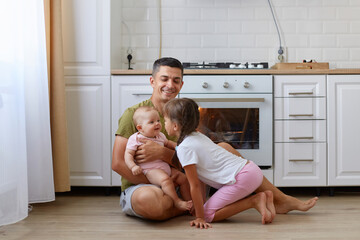 Full length portrait of happy laughing man wearing casual style t shirt sitting on floor in kitchen with daughters, cute girls playing with father, having fun, laughing.