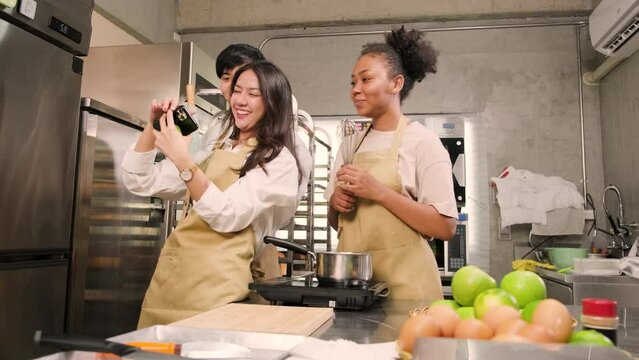 Three young students in cooking class wear aprons enjoyment while taking selfie photo with mobile phone in kitchen, smiling and laughing, preparing eggs and fruits, learn fun culinary course together.