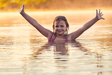 Smiling girl 10-11 years old stands in the river in summer. A happy child stands in the water and holds his hands up. Haze from the water in the early morning.