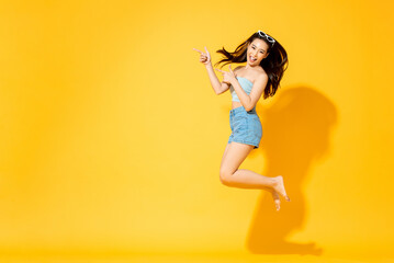 Summer portrait of smiling energetic Asian woman jumping and pointing hands to empty space in studio isolated yellow color background