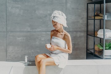 Relaxed woman applies moisturizer cream on legs after taking bath sits wrapped in towel in bathroom, enjoys beauty treatments, uses cosmetic product for healthy skin