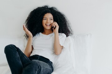Afro American woman has telephone talk, gestures and laughs with joy, looks somewhere aside, spends time at home, sits on white bed