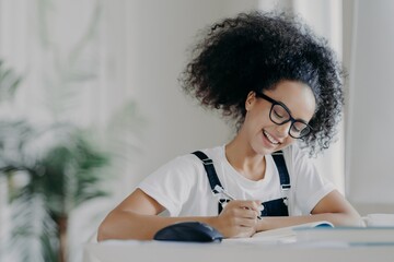 Happy Afro American female student writes down information, prepares for exams, sits in coworking space, studies in spacious room © VK Studio