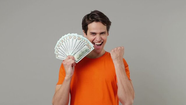 Excited satisfied joyful young brunet man 20s years old wears orange t-shirt holding fan of cash money in dollar banknotes doing winner gesture isolated on plain grey wall background studio portrait