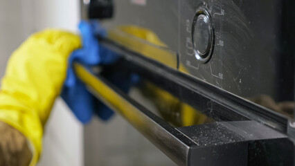 Man in yellow rubber gloves cleans oven wiping dust and stains with rag in kitchen. Housekeeper...