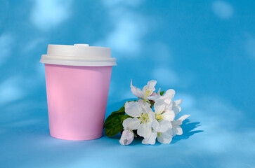 Obraz na płótnie Canvas Pink paper cup for coffee and other beverages closed with white lid, spring bouquet of apple blossoms with shadows on blue background. Natural eco packaging.