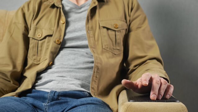 Man in mustard jacket sits in soft armchair tapping fingers on armrest against grey wall at home. Freelancer takes break after hard work closeup