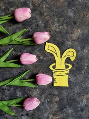 Yellow bunny ears in the hat with bouquet of pink tulips on black concrete background. Spring flatlay. Easter greeting card.