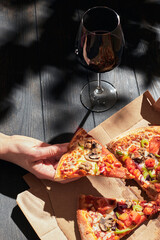 A woman's hand takes a slice of pizza on a sunny day. Pizza in a box craft and a glass of wine. The concept of food delivery. Vertical photo.