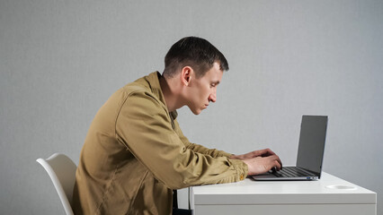 Young man works on laptop sitting at white table in office against grey wall. Focused employee in mustard jacket with crooked neck side view - obrazy, fototapety, plakaty
