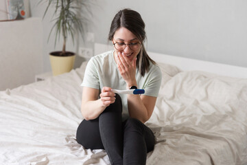 a woman looks at a pregnancy test sitting on the bed. The woman is happy and rejoices in pregnancy. A woman at home found out that she was pregnant