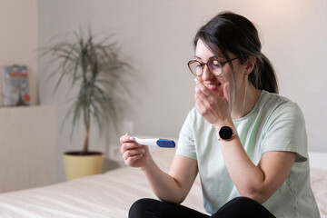 a woman looks at a pregnancy test sitting on the bed. The woman is happy and rejoices in pregnancy. A woman at home found out that she was pregnant