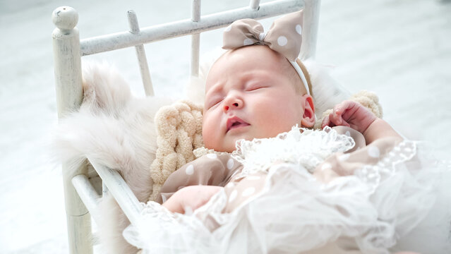Adorable newborn girl in cute dress with dotted bow on head sleeps in small cot standing on white floor. Baby photo-shoot in studio close view