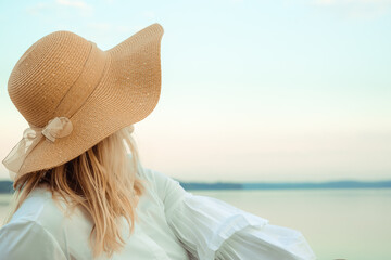 Portrait of young blond european woman in a straw hat with satin bow and white dress looking on a sea. Landscape background. Beautiful girl. Side view. Profile. Dreamed mood. International woman day