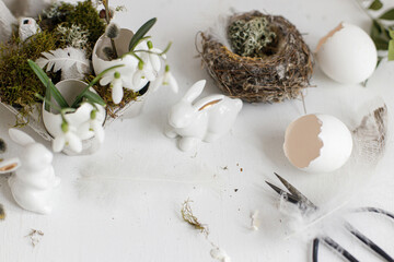 Easter rustic still life. Easter egg shells with blooming snowdrops, bunny figurines, feathers, nest on aged white wooden table. Simple stylish festive decoration on table. Happy Easter!