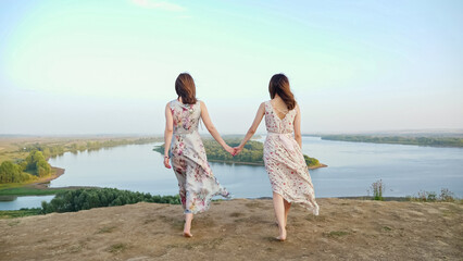 Graceful brunette barefoot women in long dresses walk joining hands along hilly riverbank at...