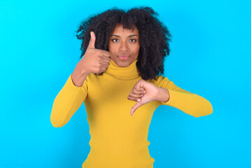 Young woman with afro hairstyle wearing yellow turtleneck over blue background showing thumbs up...