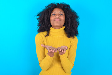 Young woman with afro hairstyle wearing yellow turtleneck over blue background holding something...