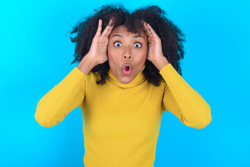 Young woman with afro hairstyle wearing yellow turtleneck over blue background with scared...