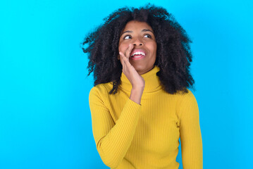 Fototapeta premium Young woman with afro hairstyle wearing yellow turtleneck over blue background hear incredible private news impressed scream share