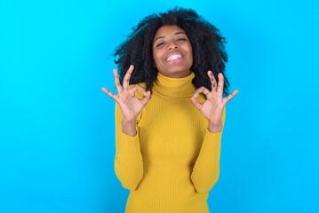 Young woman with afro hairstyle wearing yellow turtleneck over blue background showing both hands...